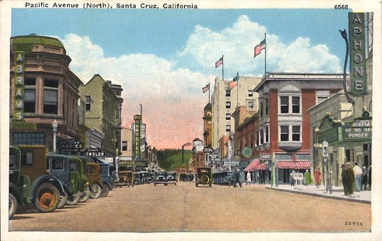 Pacific Avenue in Santa Cruz, 1930. The Bank of Italy is the green building on the right at the corner of Soquel and Pacific. The distinctive red brick Trust Building is the street. In the distance, the Palomar and IOOF building with its clock tower. On the left, the Hihn building, and the New Santa Cruz Theatre neon in the distance. Image: Linda Rosewood postcard collection